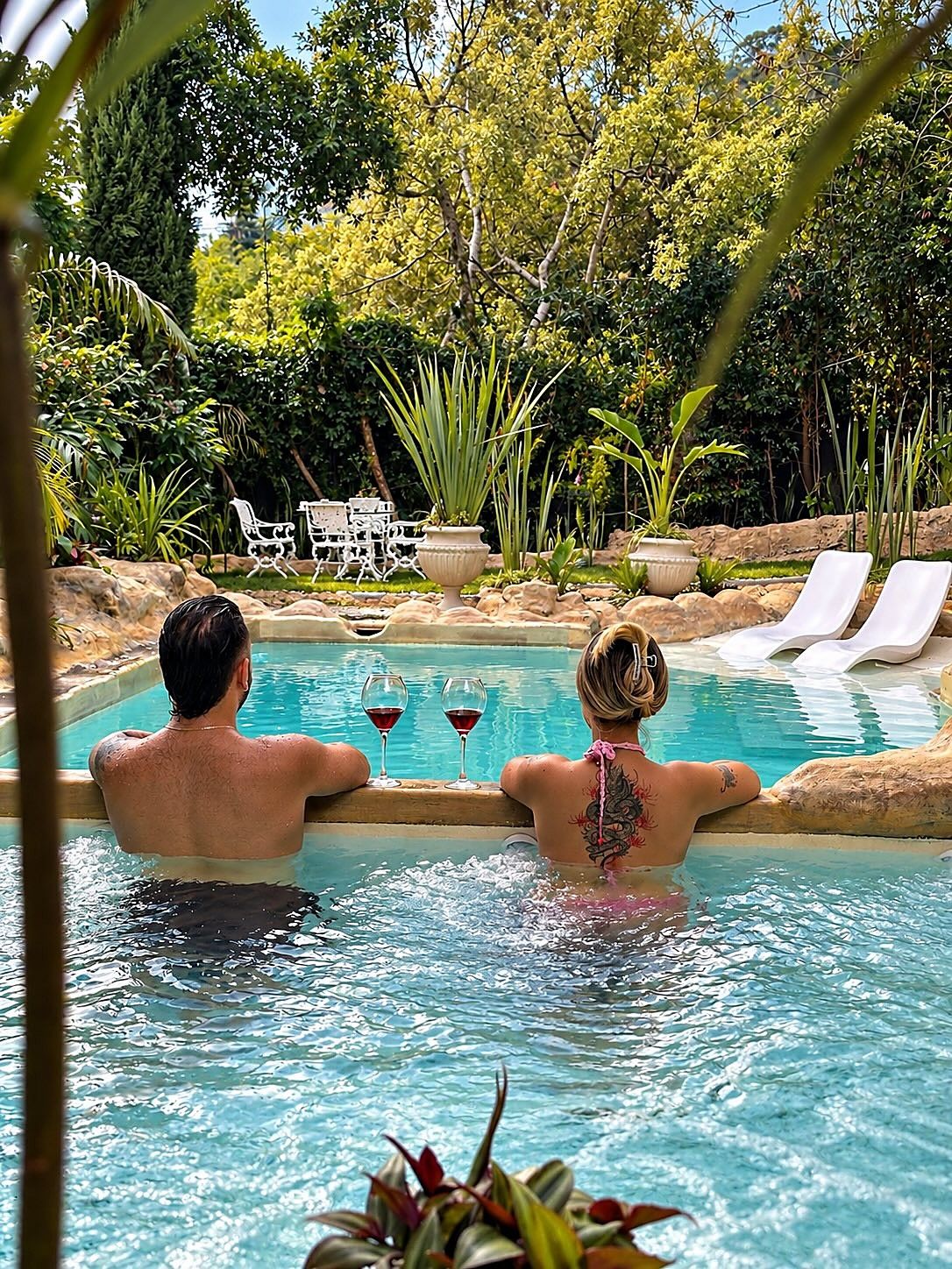 Pareja de espaldas dentro de la piscina de Garden SPA, cada uno con una copa de vino, entre rocas y jardín