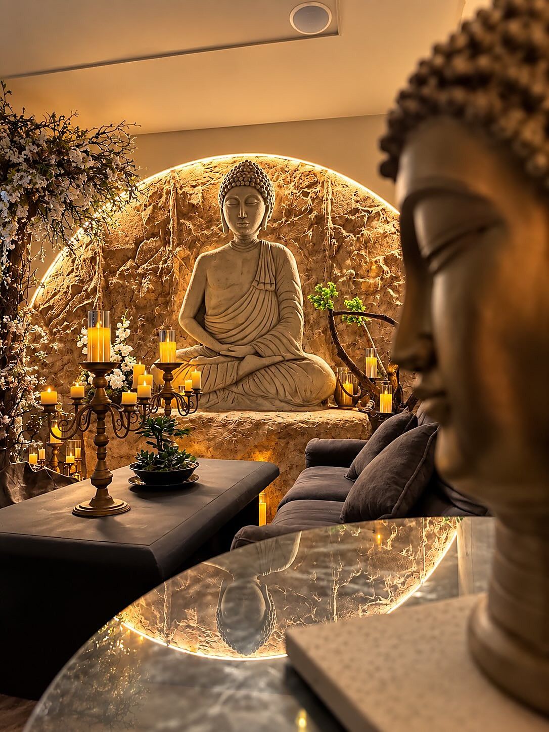 Cabeza de Buda en bronce en primer plano y, al fondo, un Buda tallado en piedra dentro de un círculo iluminado, con velas encendidas